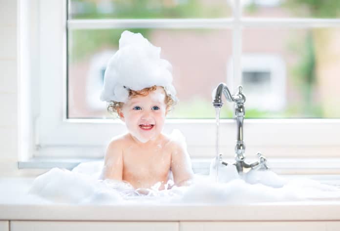 Playing with water drops and splashes next to a big window with garden view funny little baby girl with wet curly hair taking a bath in a kitchen sink with lots of foam, kid with bubble bath in bathtub